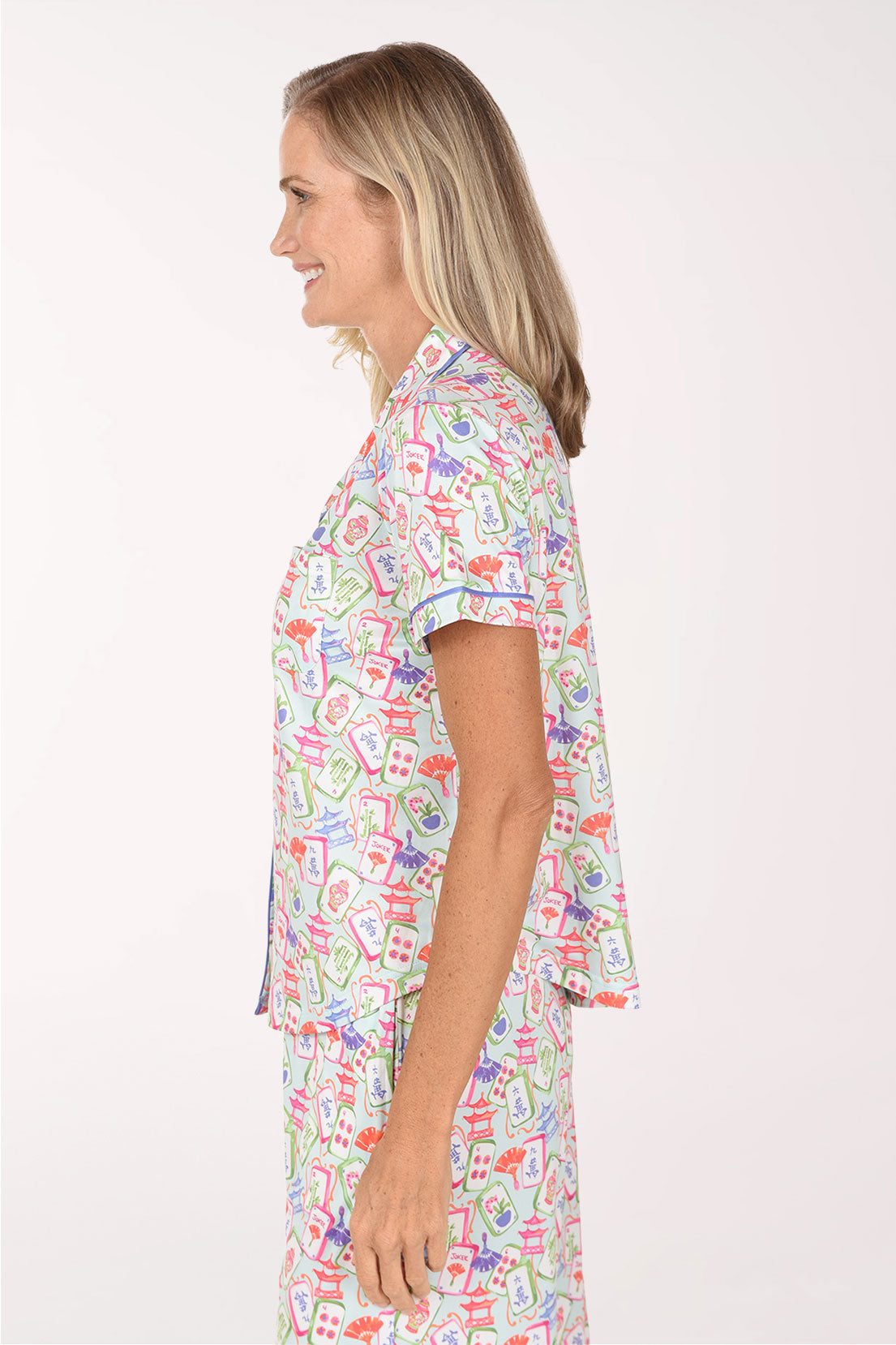 A smiling blonde woman wearing a short-sleeve button-front pajama top and matching pants with a colorful cartoon-style print on a white base, photographed against a plain light background.