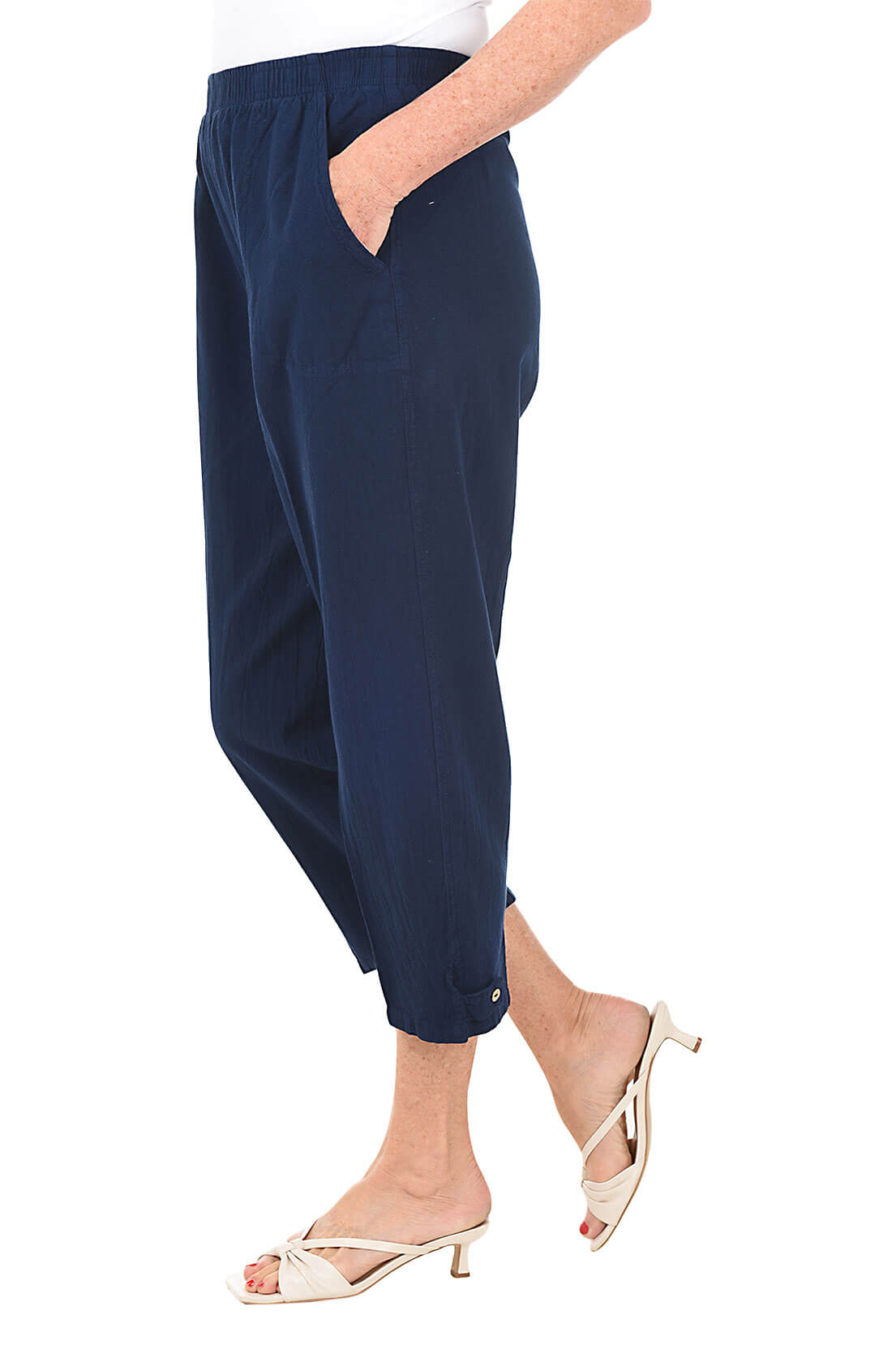 A side shot of a model wearing a pair of navy blue capri pants with button-tab detail on the hemline, made of crinkle cotton fabric, with an elastic waistband and side pockets.