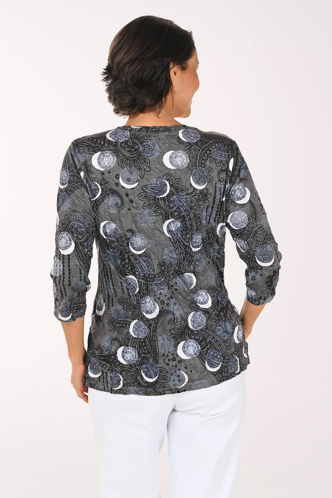 A smiling woman wearing a dark gray wrap-style blouse with a moon phase pattern and white pants, photographed against a plain white studio background.