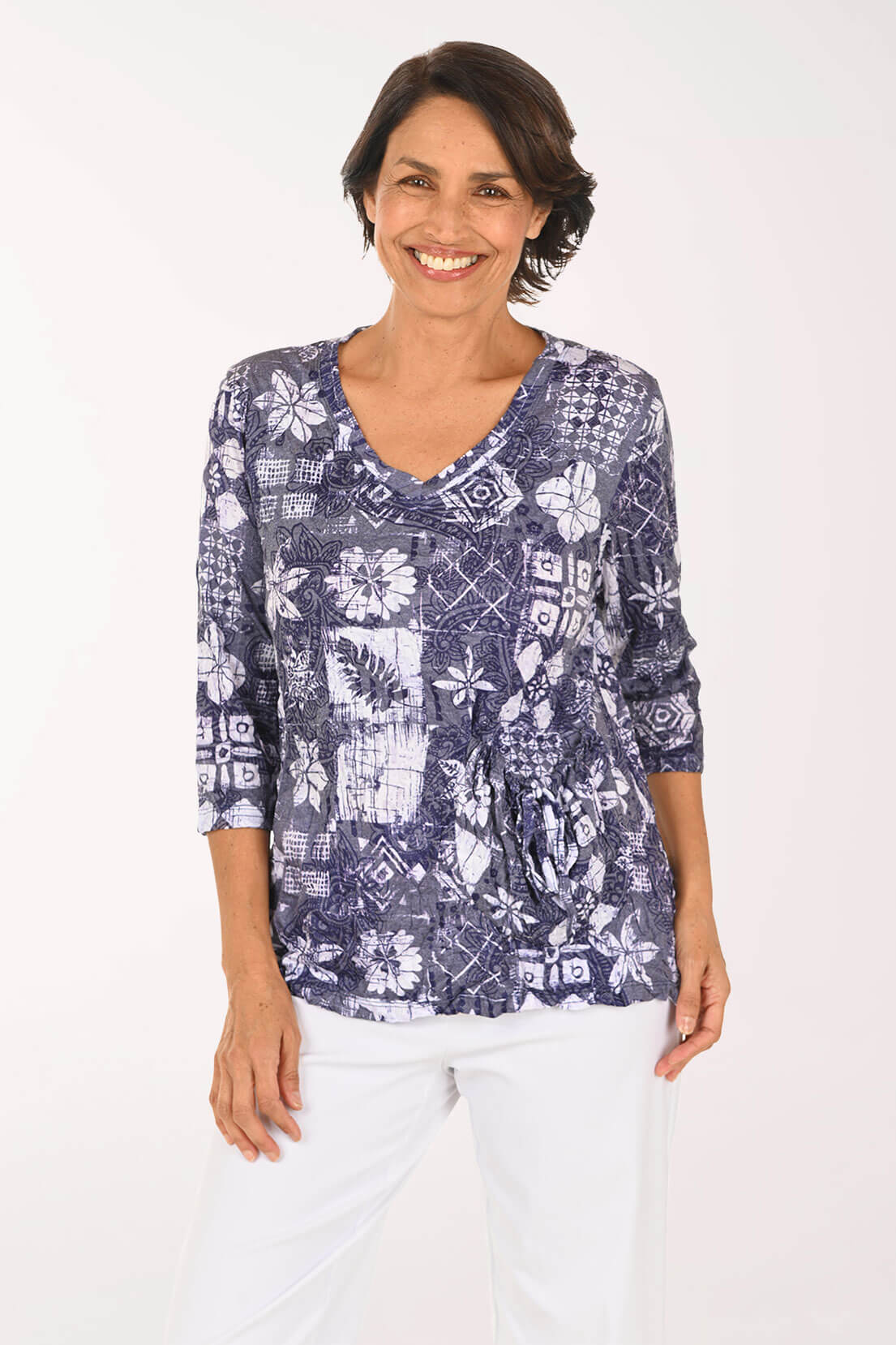A smiling woman wearing a blue and white patterned three-quarter sleeve top with white pants, photographed against a plain light background.
