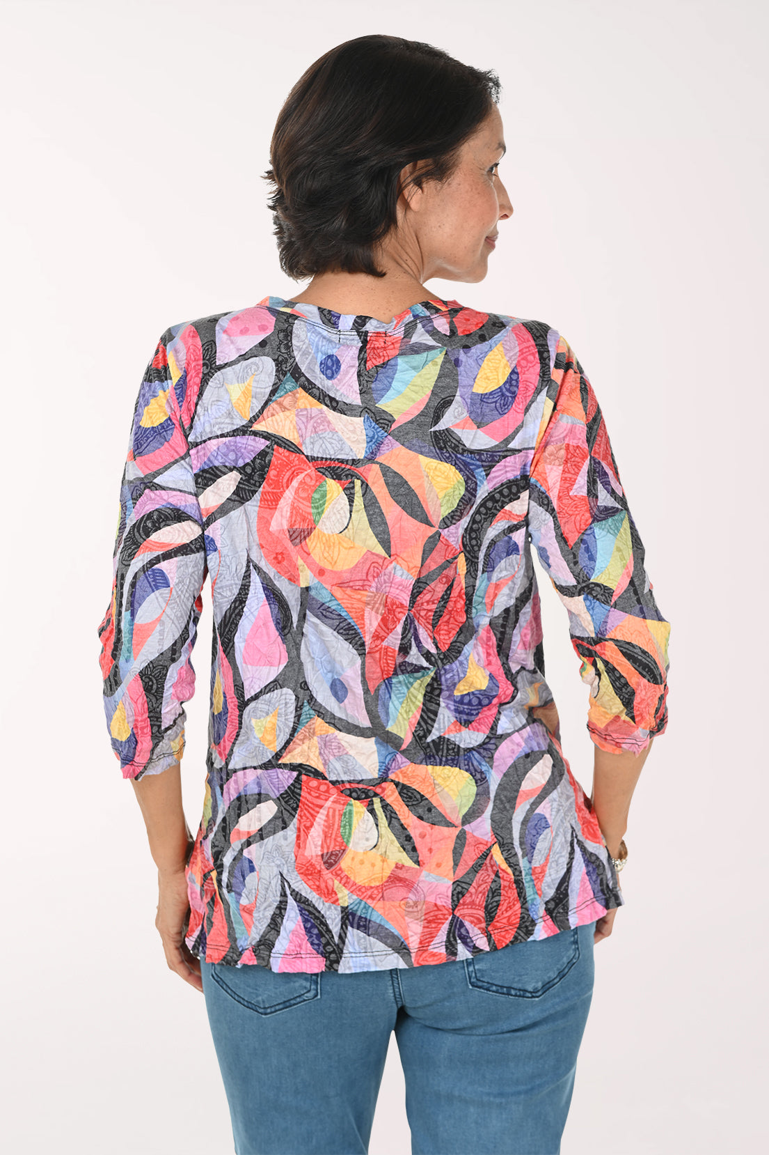 A smiling woman wearing a colorful abstract print top with three-quarter sleeves and blue jeans, standing against a plain white studio background.
