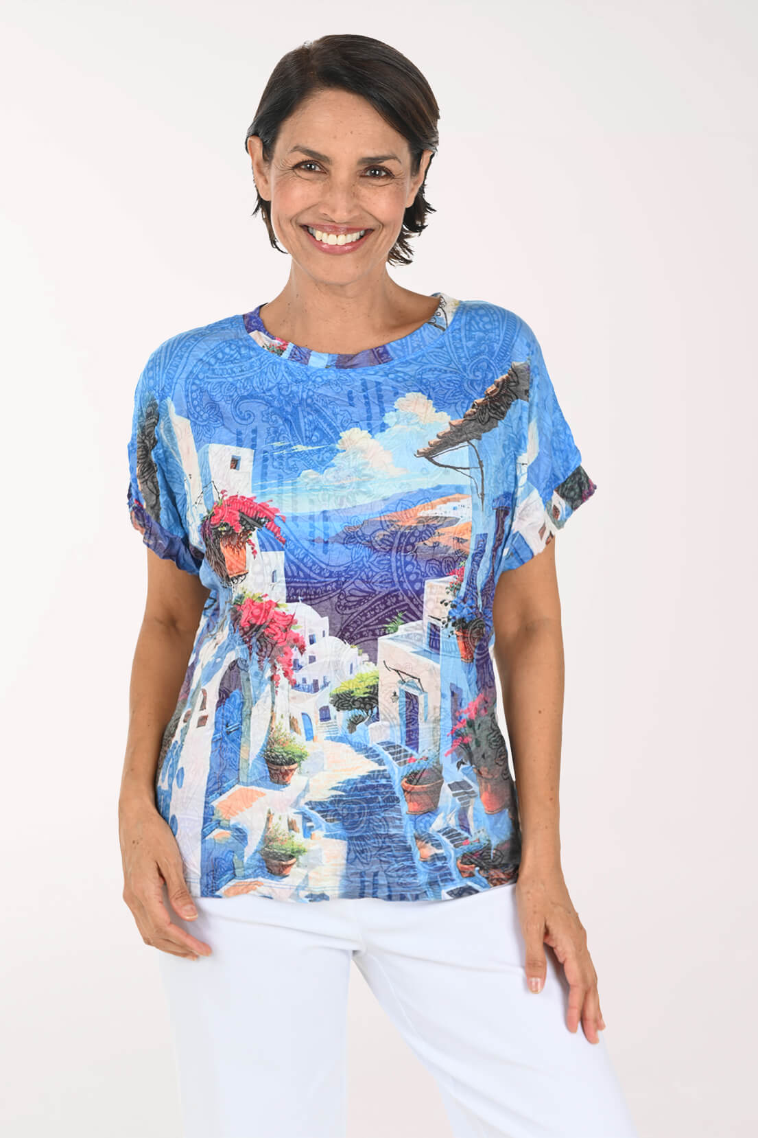 A woman wearing a short-sleeve blue top with a scenic Mediterranean village print, paired with white pants, against a plain white background.
