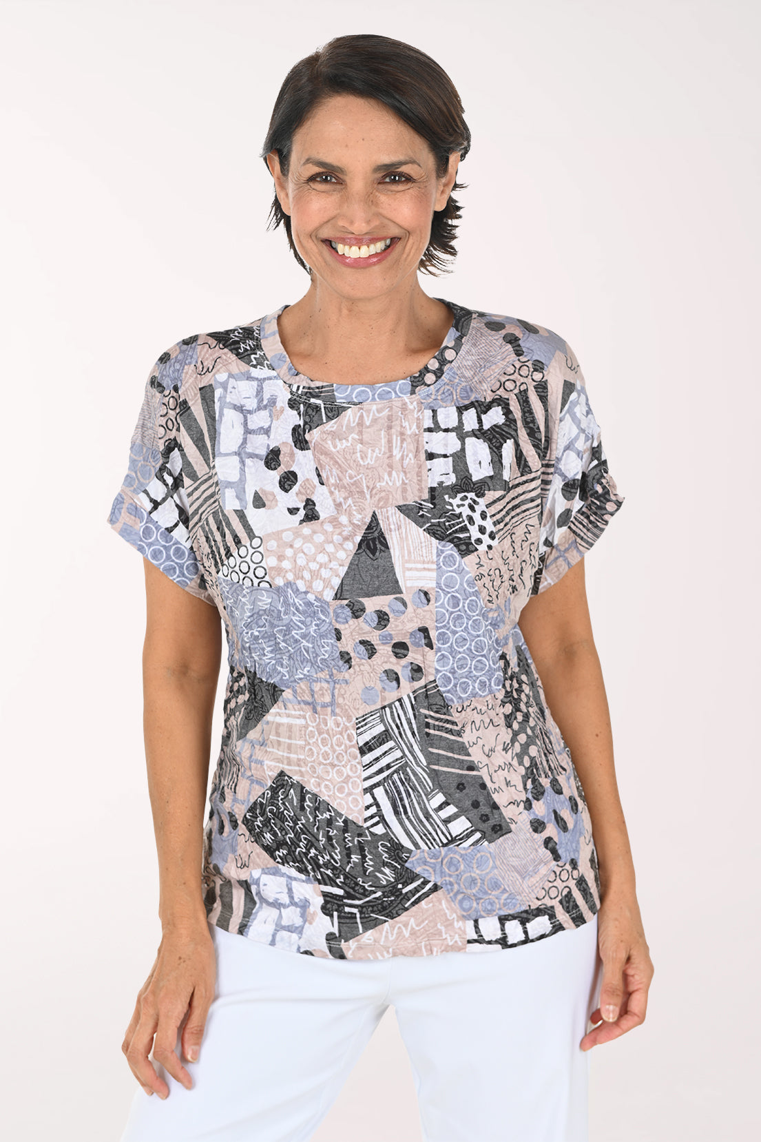 A smiling woman wearing a short-sleeve blouse with a multicolor abstract patchwork print in shades of blue, gray, black, and beige, paired with white pants, against a plain white studio background.