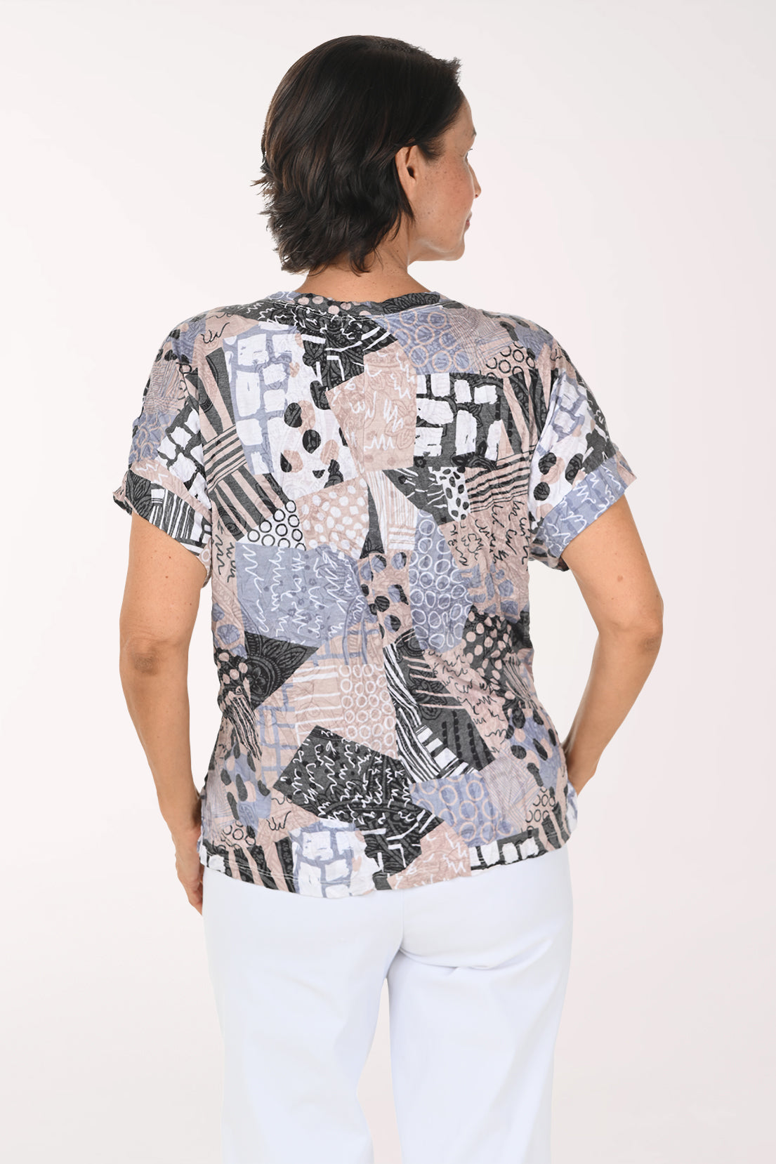 A smiling woman wearing a short-sleeve blouse with a multicolor abstract patchwork print in shades of blue, gray, black, and beige, paired with white pants, against a plain white studio background.