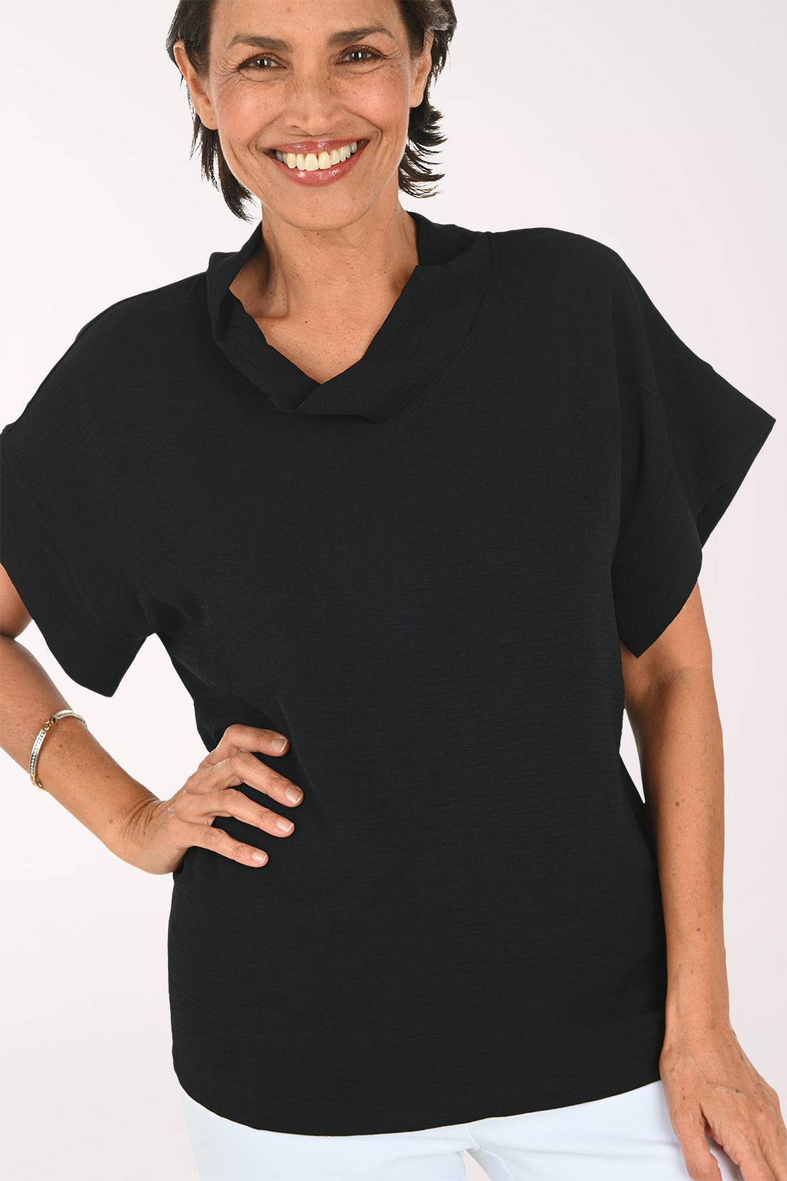 A smiling woman wearing a black short-sleeve cowl-neck top and white pants, standing against a plain white studio background.