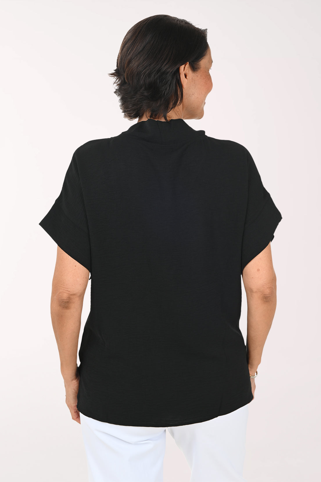 A smiling woman wearing a black short-sleeve cowl-neck top and white pants, standing against a plain white studio background.