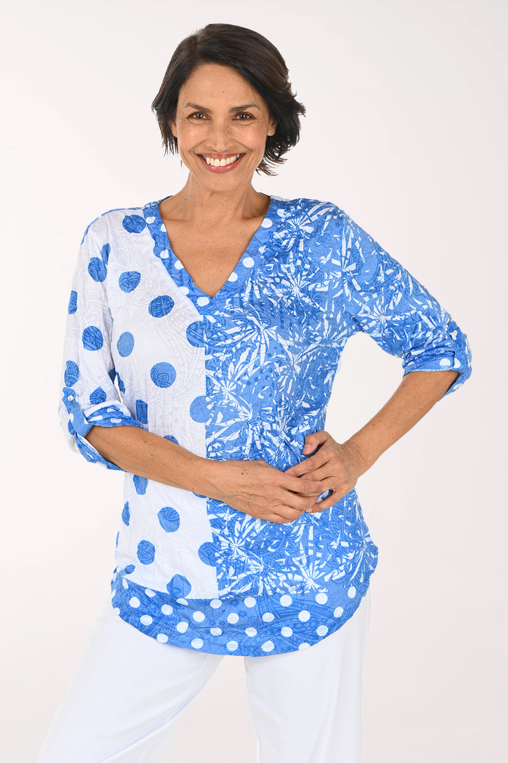 A woman modeling a blue and white patterned top with three-quarter sleeves and a V-neck, paired with white pants, against a plain white studio background.