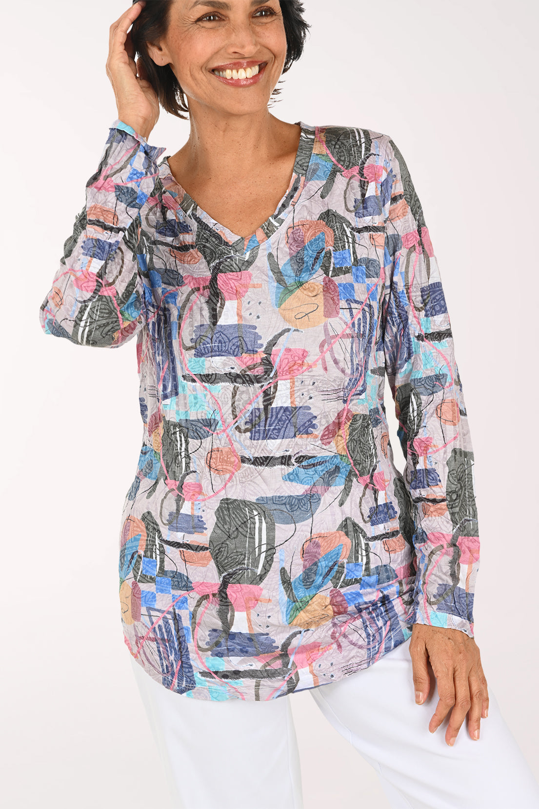 A smiling woman with short brown hair stands against a plain white background wearing a long-sleeve V-neck top with a colorful abstract print and white pants.