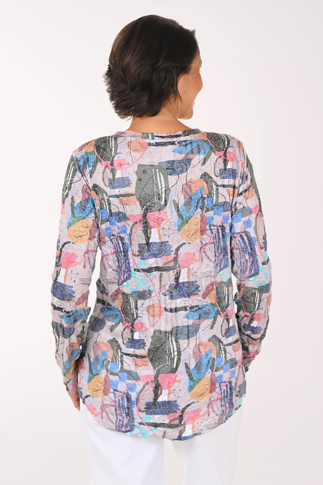 A smiling woman with short brown hair stands against a plain white background wearing a long-sleeve V-neck top with a colorful abstract print and white pants.
