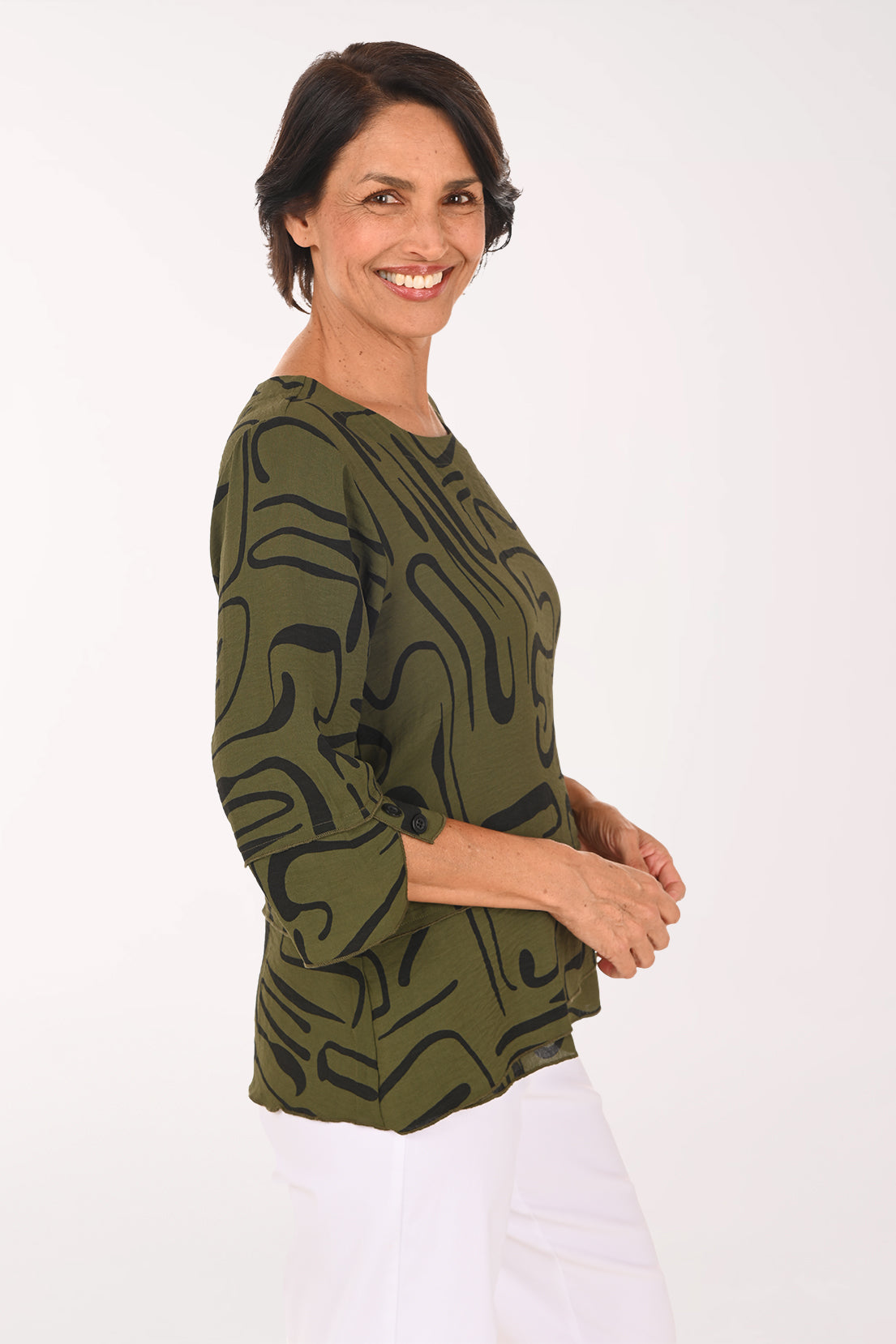 A smiling woman wearing an olive green and black abstract patterned blouse with three-quarter ruffled sleeves, paired with white pants, against a plain white studio background.