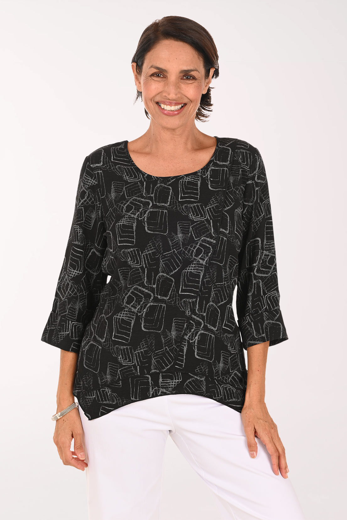 A woman wearing a black three-quarter sleeve top with a white geometric line pattern, paired with white pants, standing against a plain white background.
