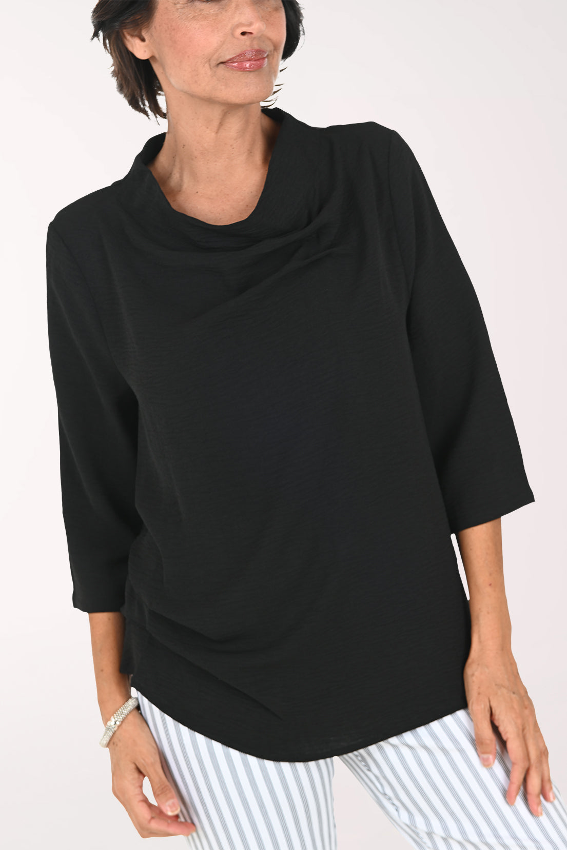 A smiling woman with short brown hair wearing a black cowl-neck three-quarter sleeve top and white and gray vertically striped pants, posing against a plain white studio background.
