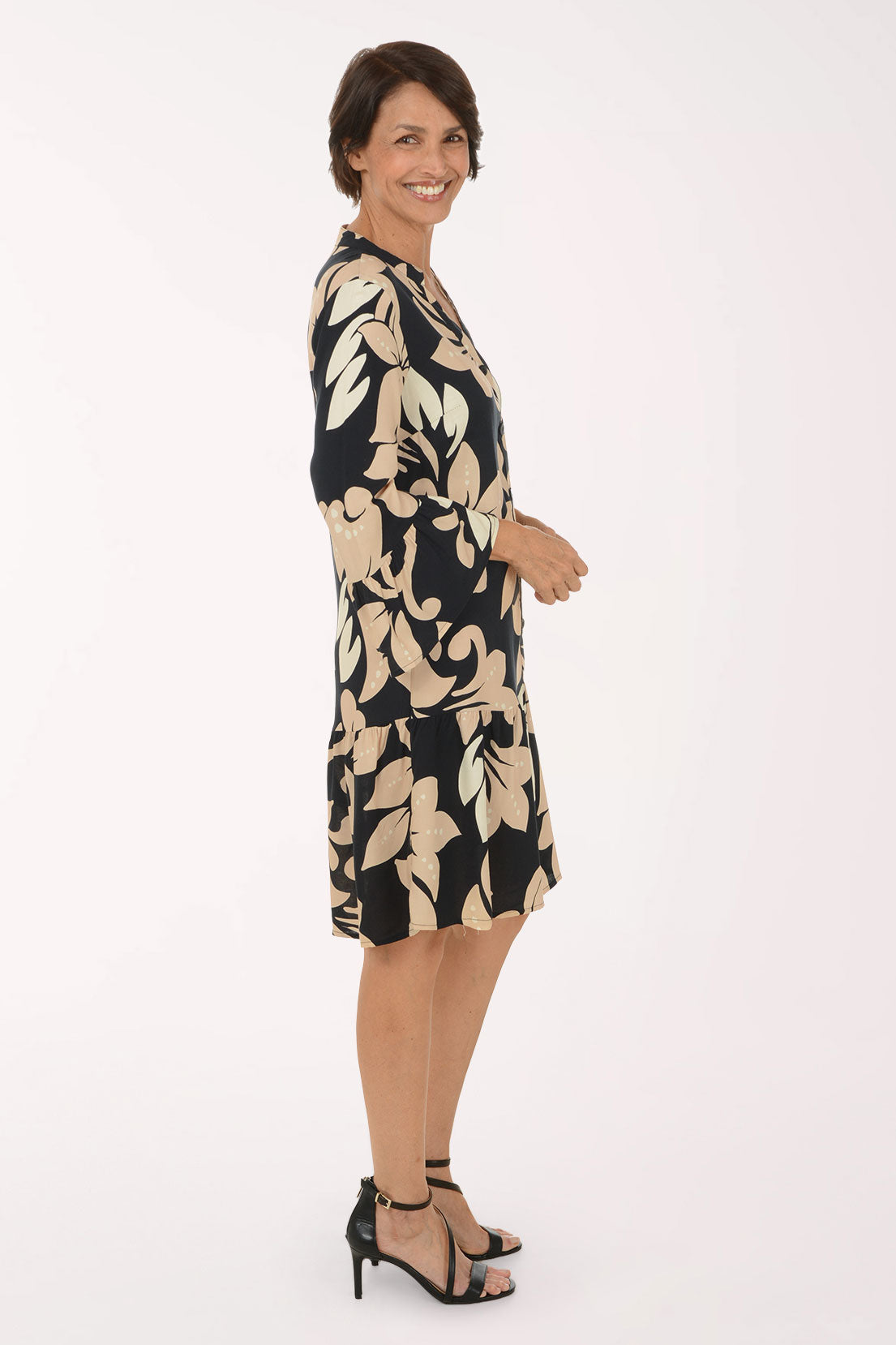 A woman modeling a knee-length black and beige floral print dress with three-quarter bell sleeves and a button-up front, paired with black strappy high-heeled sandals, against a plain white studio background.