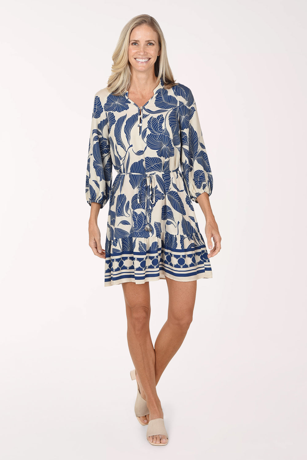 Model wearing the Mlle Gabrielle Trumpet Flower Belted Dress, a cream dress with royal blue trumpet florals and a geometric-border ruffle hem, front view