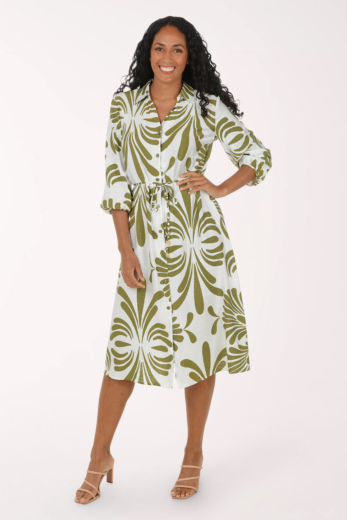 A woman modeling a white midi shirt dress with a green palm leaf pattern, featuring three-quarter sleeves, a button-up front, and a matching tie belt at the waist, paired with beige strappy heels against a solid white studio background.
