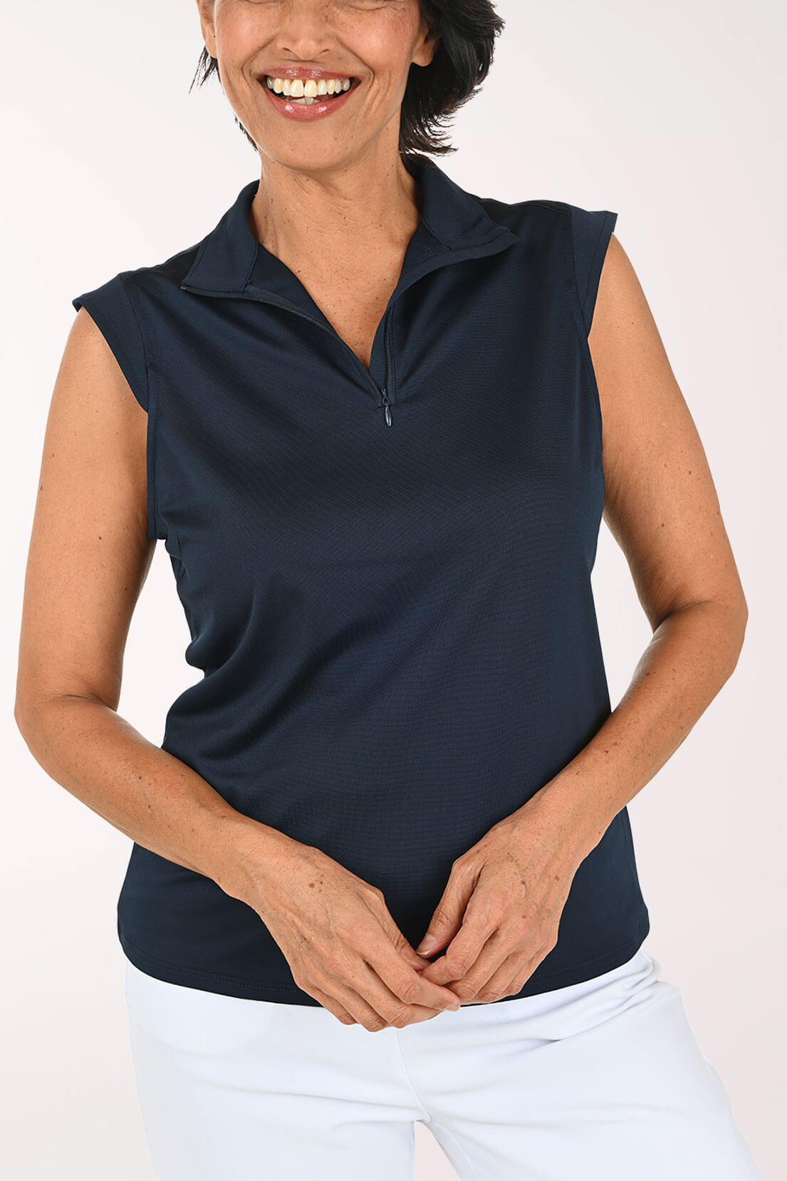 A woman wearing a sleeveless dark navy polo shirt with a short zipper at the collar, paired with white pants, against a plain light background.