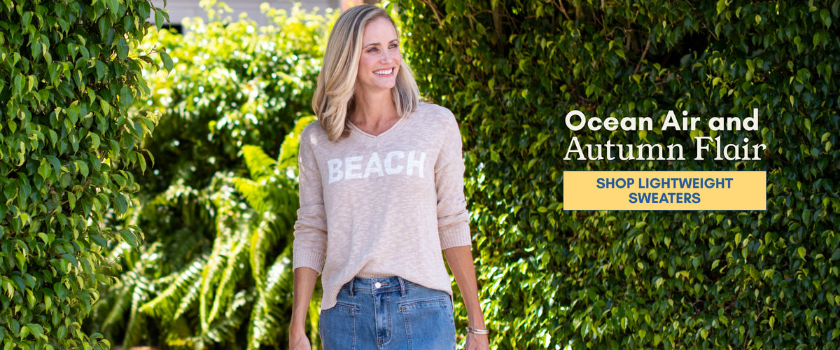 Woman wearing a lightweight beige BEACH sweater with jeans outdoors surrounded by lush greenery.