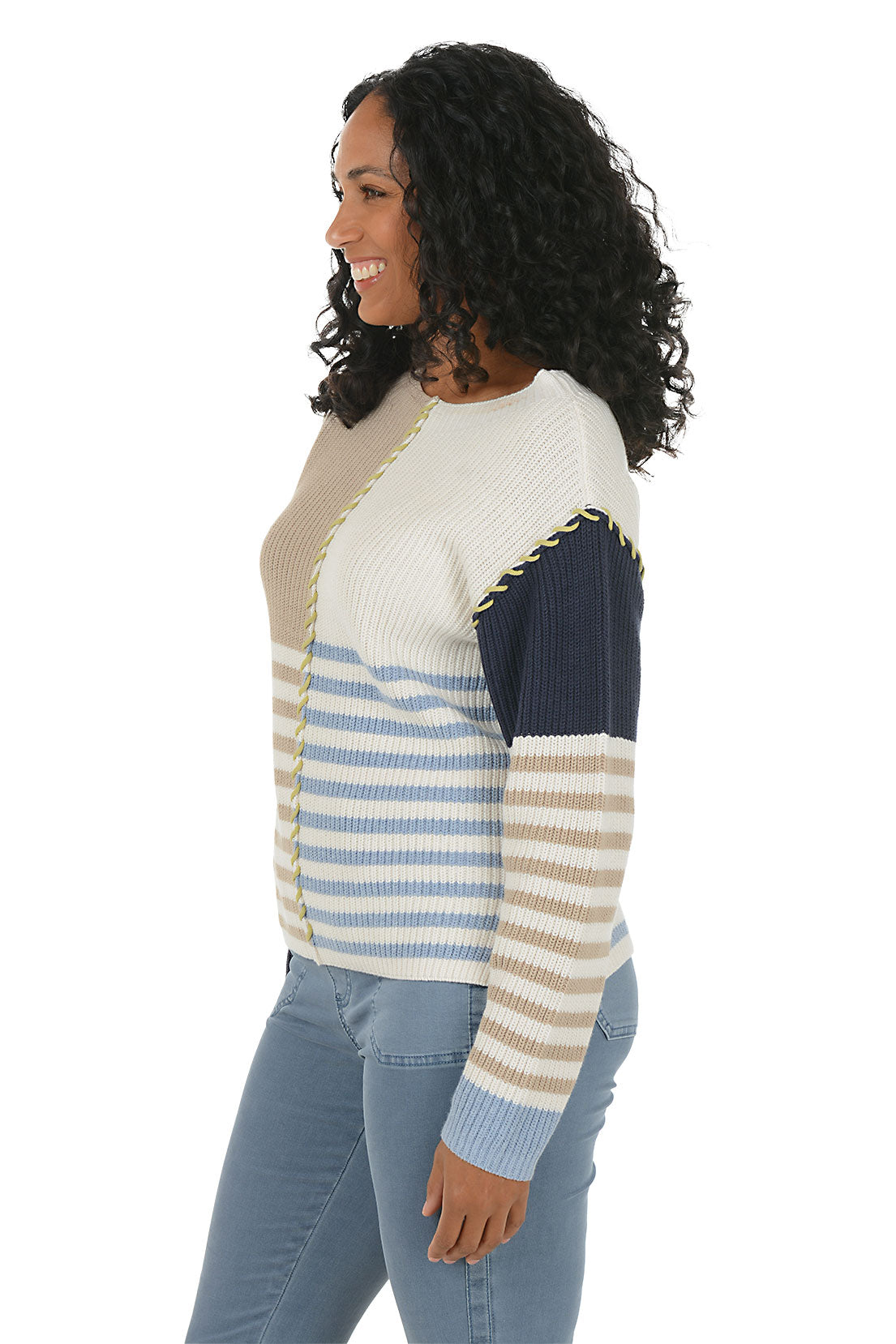 Woman wearing a colorful patchwork sweater boat neck sweater with blue jeans on a white background. Side shot.