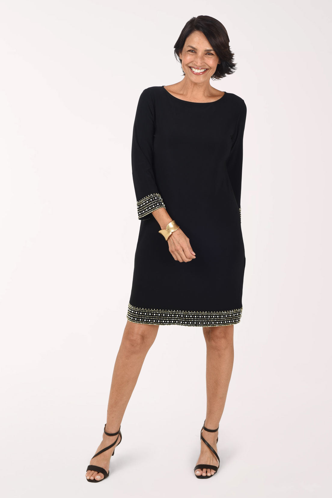 Front view of the black Tiana B Beaded Trim 3/4 Sleeve Dress, a straight above-the-knee shift with a boat neckline and wide bands of gold beads and pearls at the sleeve cuffs and hem, worn with a gold cuff bracelet and black strappy heels