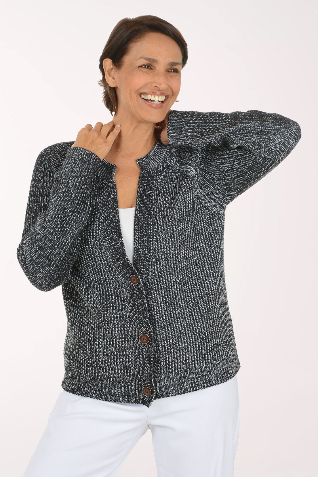A smiling woman wearing a dark gray and black marled knit cardigan with brown buttons over a white top and white pants, photographed against a plain light background.