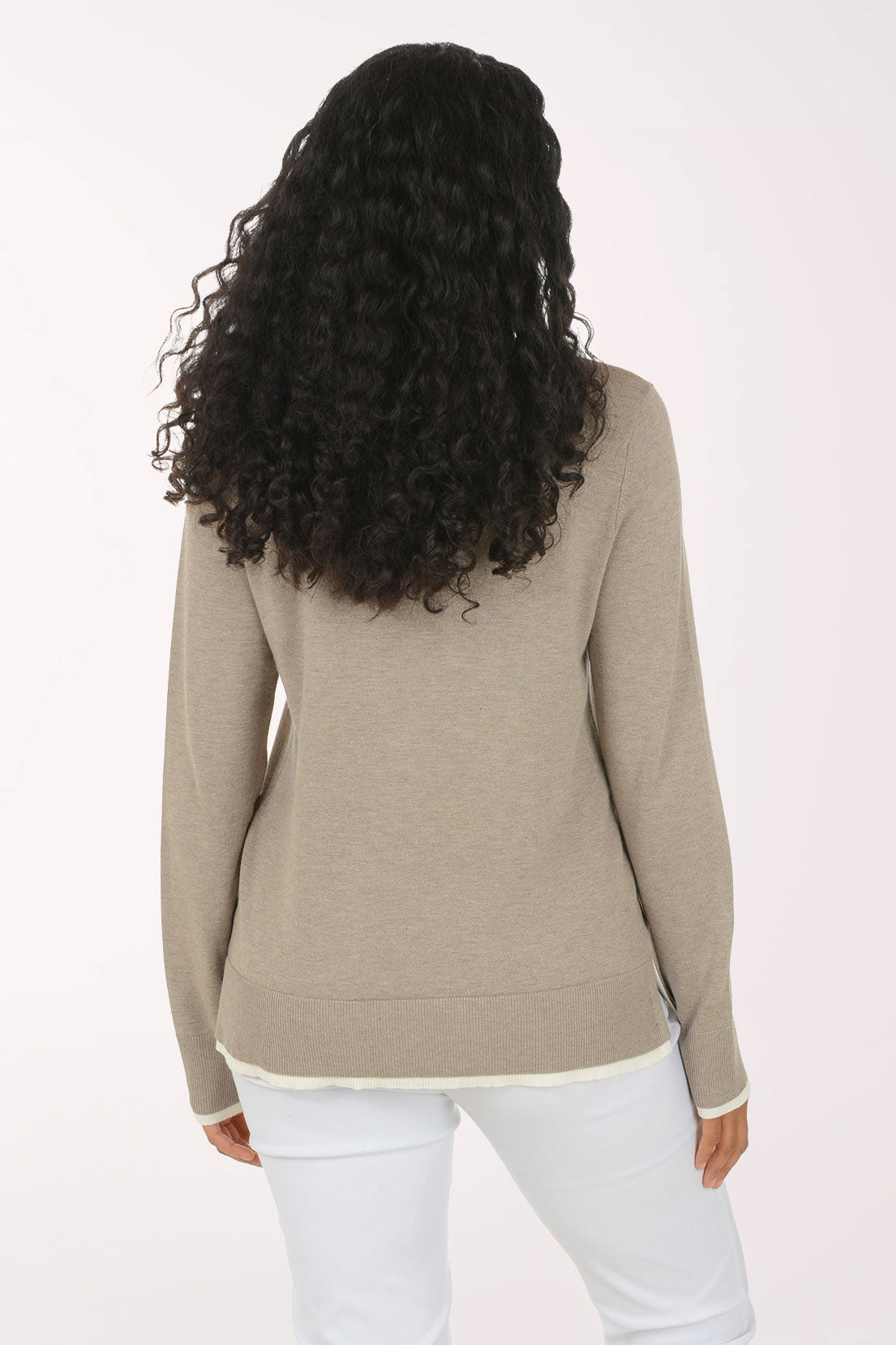 A smiling woman with long curly dark hair wears a taupe-beige long-sleeve V-neck sweater with white contrast trim at the neckline, cuffs, and hem, paired with white pants, against a plain light background.