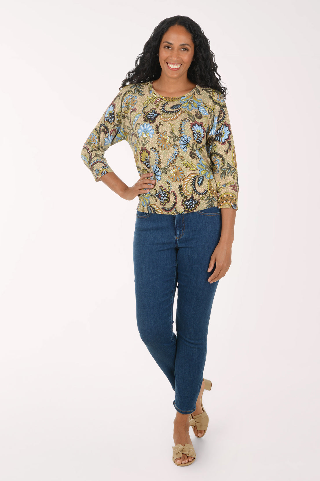 A woman modeling a three-quarter sleeve top with a detailed gold, blue, and brown paisley pattern, paired with dark blue skinny jeans and beige flat sandals, against a plain white studio background.