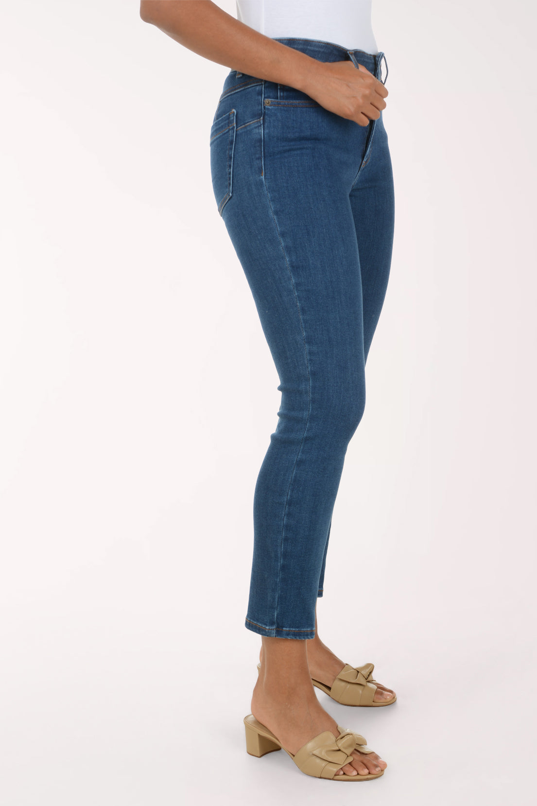 A person wearing blue skinny jeans with a side slit at the hem, paired with a white top and beige slide sandals, shown from the waist down against a plain white studio background.