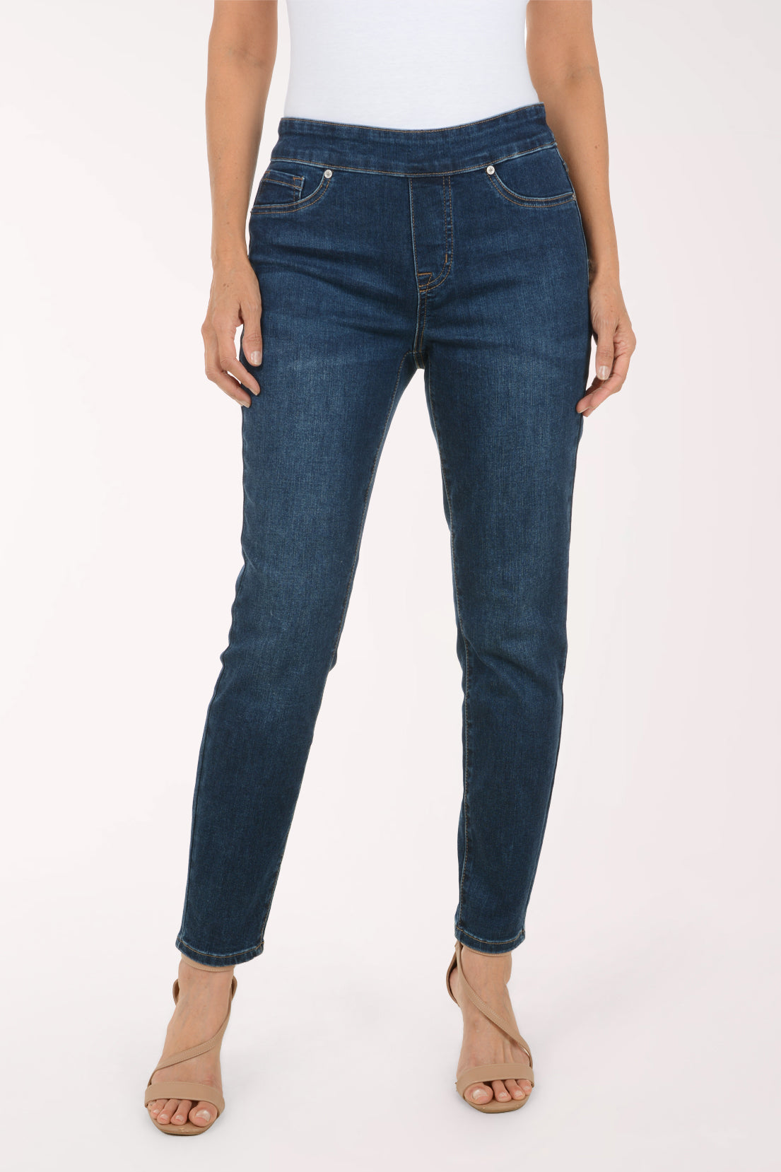 A model wearing dark blue skinny jeans with a white top and beige heeled sandals, shown from the waist down against a plain white studio background.