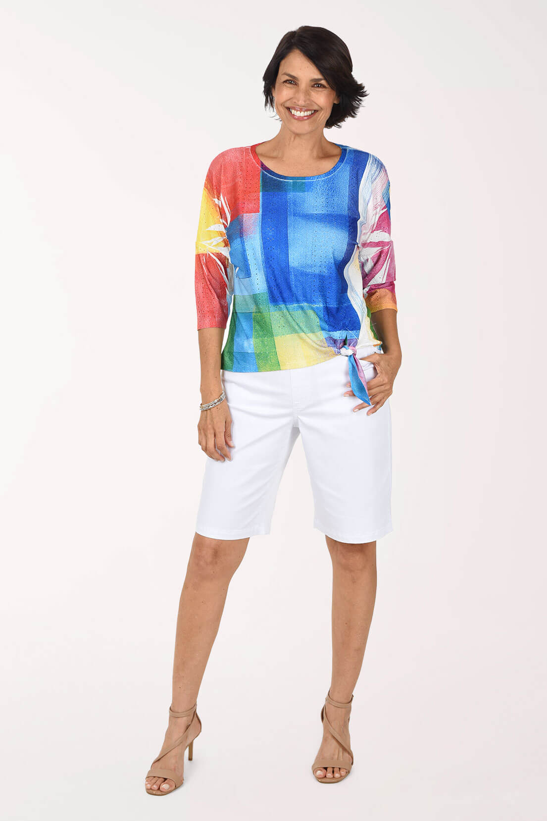 Full-length front view of a woman in white stretch denim Bermuda shorts worn with a bright multicolor color-block print three-quarter sleeve top tied at the hip and nude strappy heeled sandals.