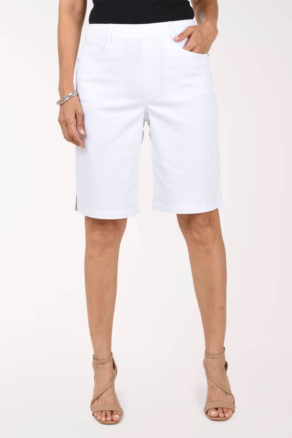 Front view of white stretch denim Bermuda shorts with a pull-on waistband, belt loops, five-pocket styling, faux fly and small side slits at the hem, worn with a black top and nude sandals.