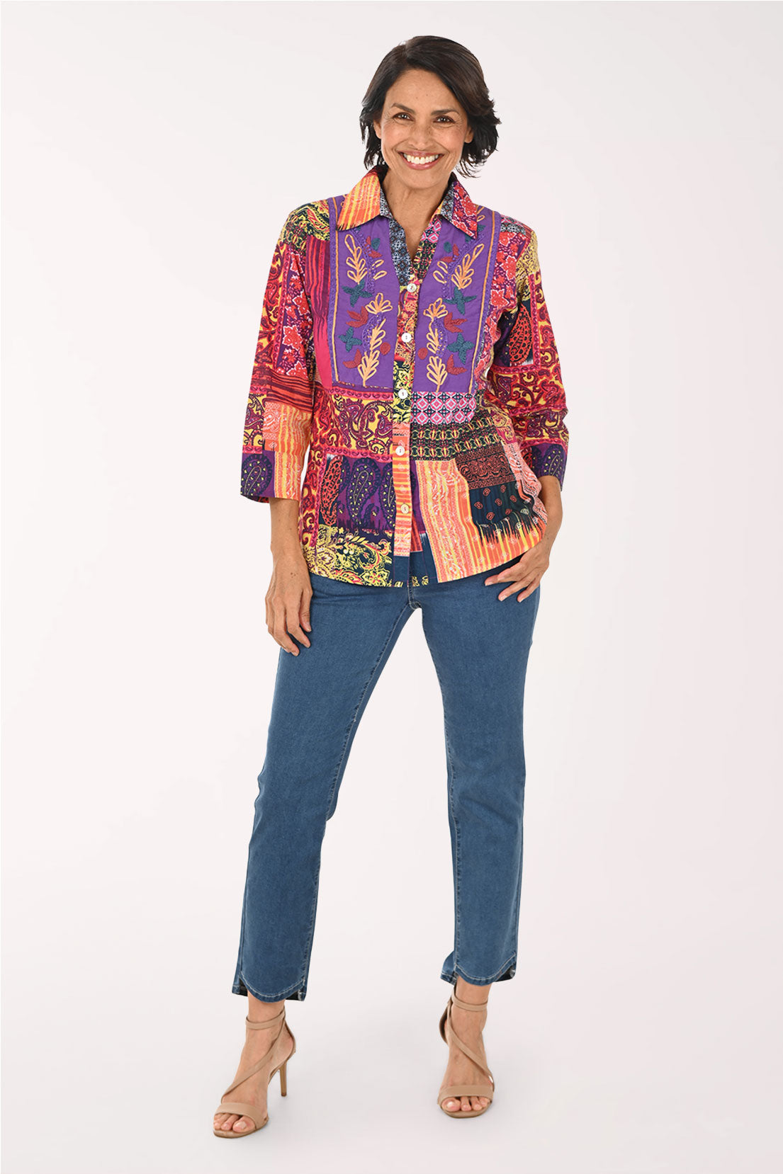 A woman modeling a vibrant, multicolored patchwork button-up shirt with blue jeans and beige high-heeled sandals, standing against a plain white studio background.