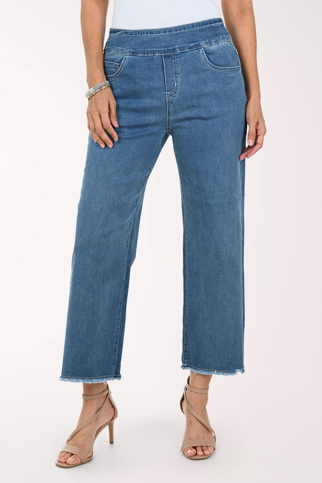 A model wearing medium-wash blue cropped jeans with a raw frayed hem, paired with beige strappy high-heeled sandals against a plain light background.
