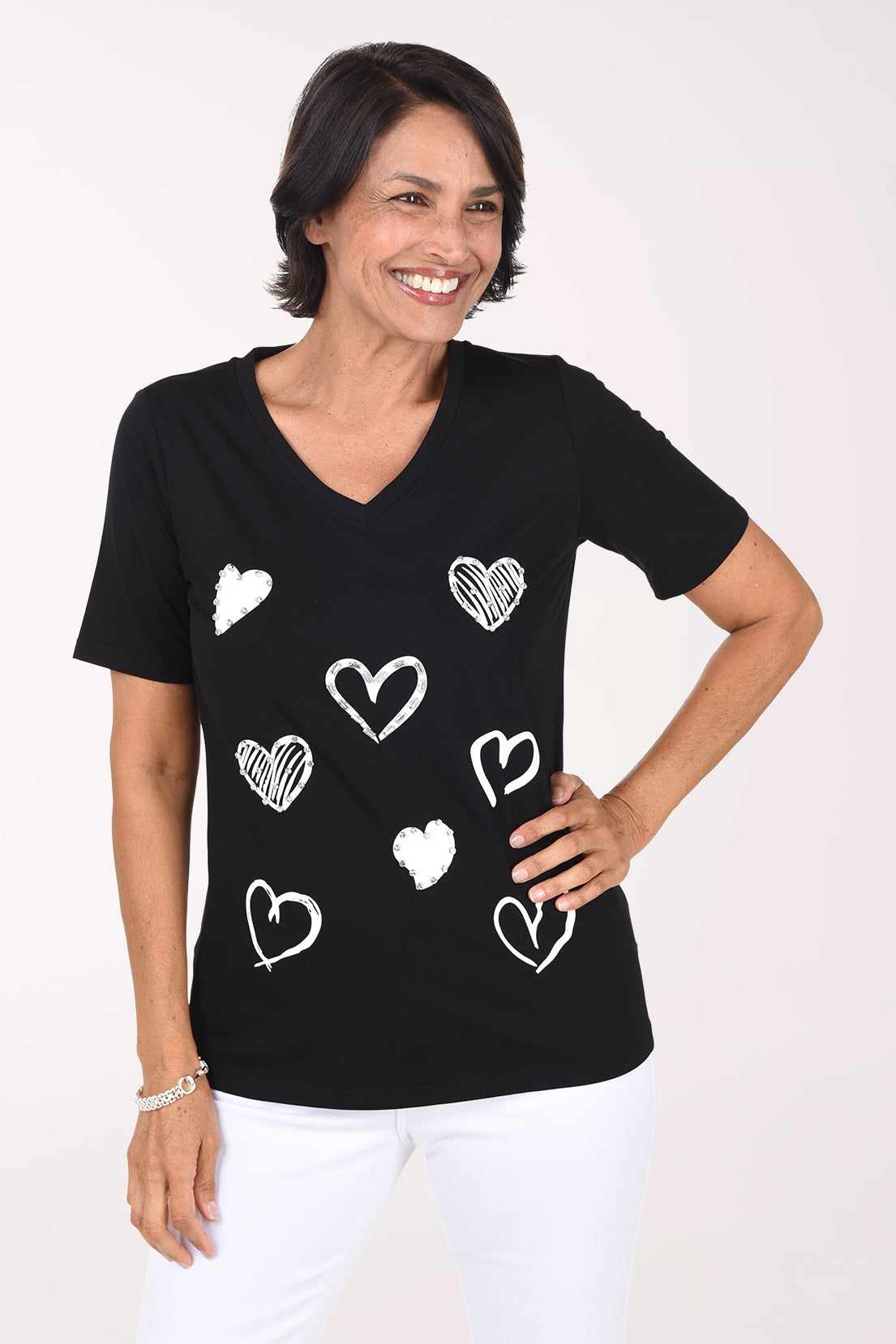 Front view of the black V-neck tee with scattered sketched hearts, worn with white pants, one hand on the hip.