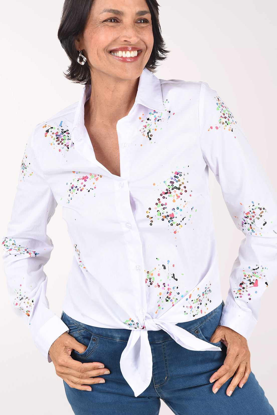 Front close-up of the white shirt showing the pink, green, blue and gold paint speckles and sequins, the button placket and the tied hem.