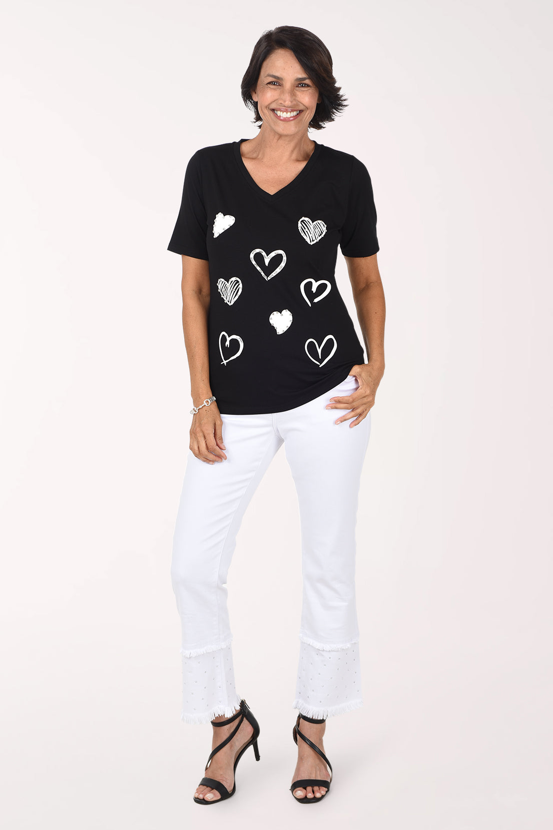 Front view of a woman in white stretch denim ankle-length flared pants with a double fringed hem, the lower tier scattered with silver rhinestones, worn with a black V-neck tee with white hearts and black strappy heeled sandals.
