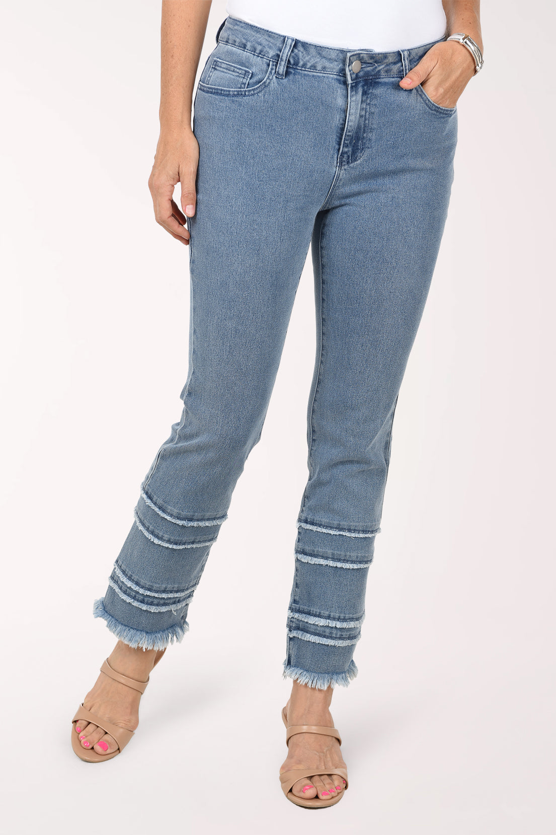 Front view of light-wash stretch denim straight leg ankle pants with several frayed tiers stacked at the hem, worn with a white top, one hand in the front pocket and nude sandals.