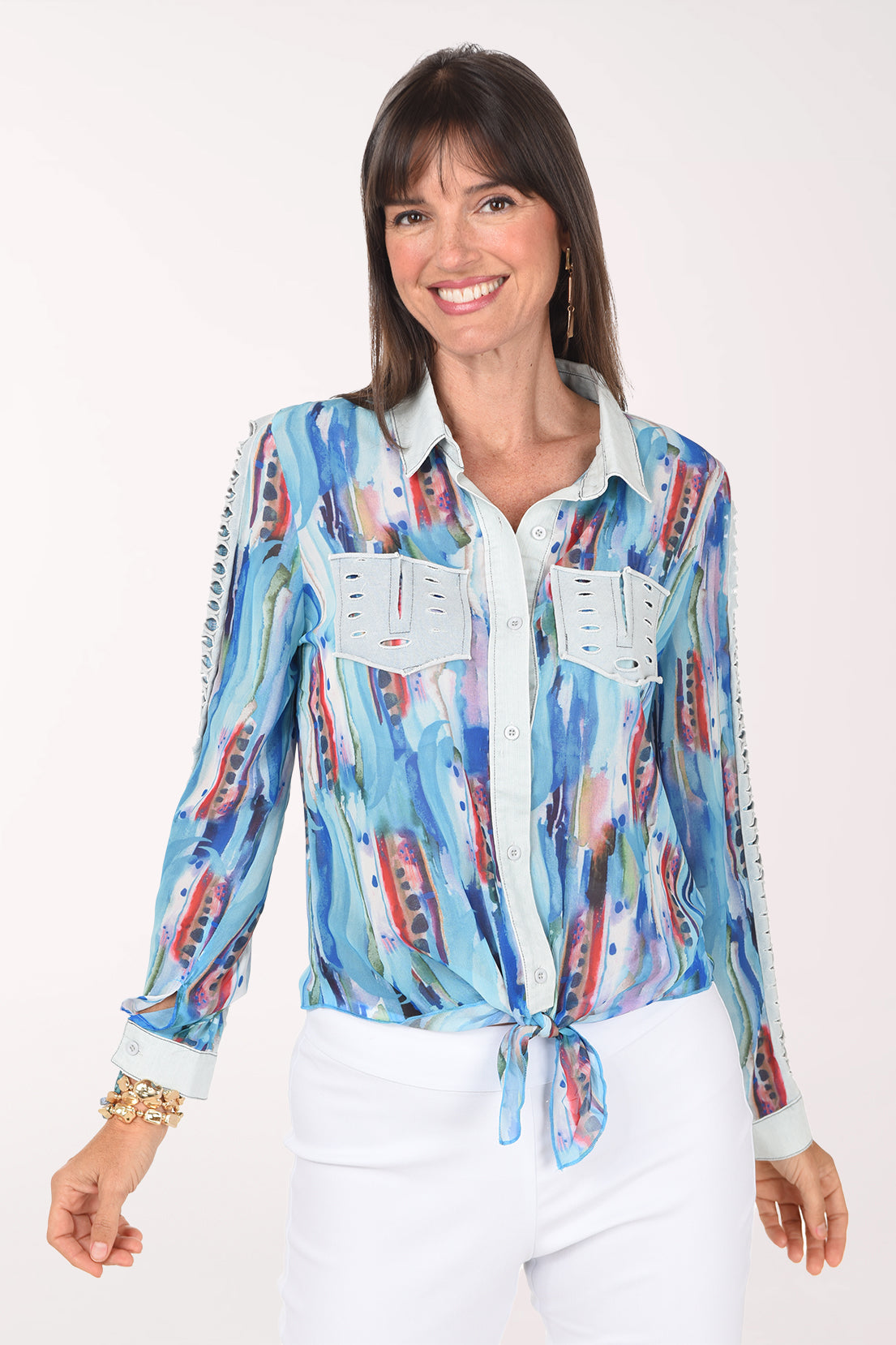 Front view of a woman in a long sleeve chiffon shirt with a turquoise, cobalt and sky blue watercolor brushstroke print with touches of red, light denim collar, placket and faux pockets, tied at the hem over white pants.