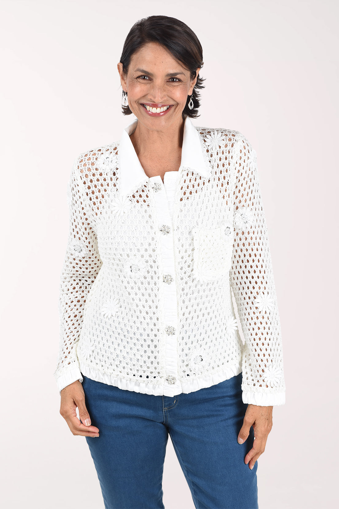 Front view of a woman in an ivory open crochet long sleeve jacket with a white satin collar, placket, cuffs and hem, jeweled floral buttons, a chest pocket and floral appliques, worn over a white cami with jeans.