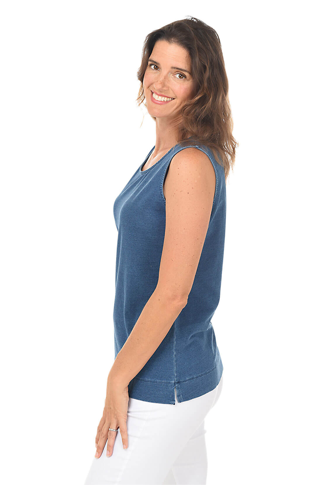 Woman wearing a blue sleeveless top and white pants on a white background. Side shot.