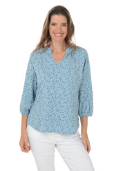 A front shot of a woman wearing a blue dotted split neck slub knit top with 3/4 raglan sleeves and white pants.