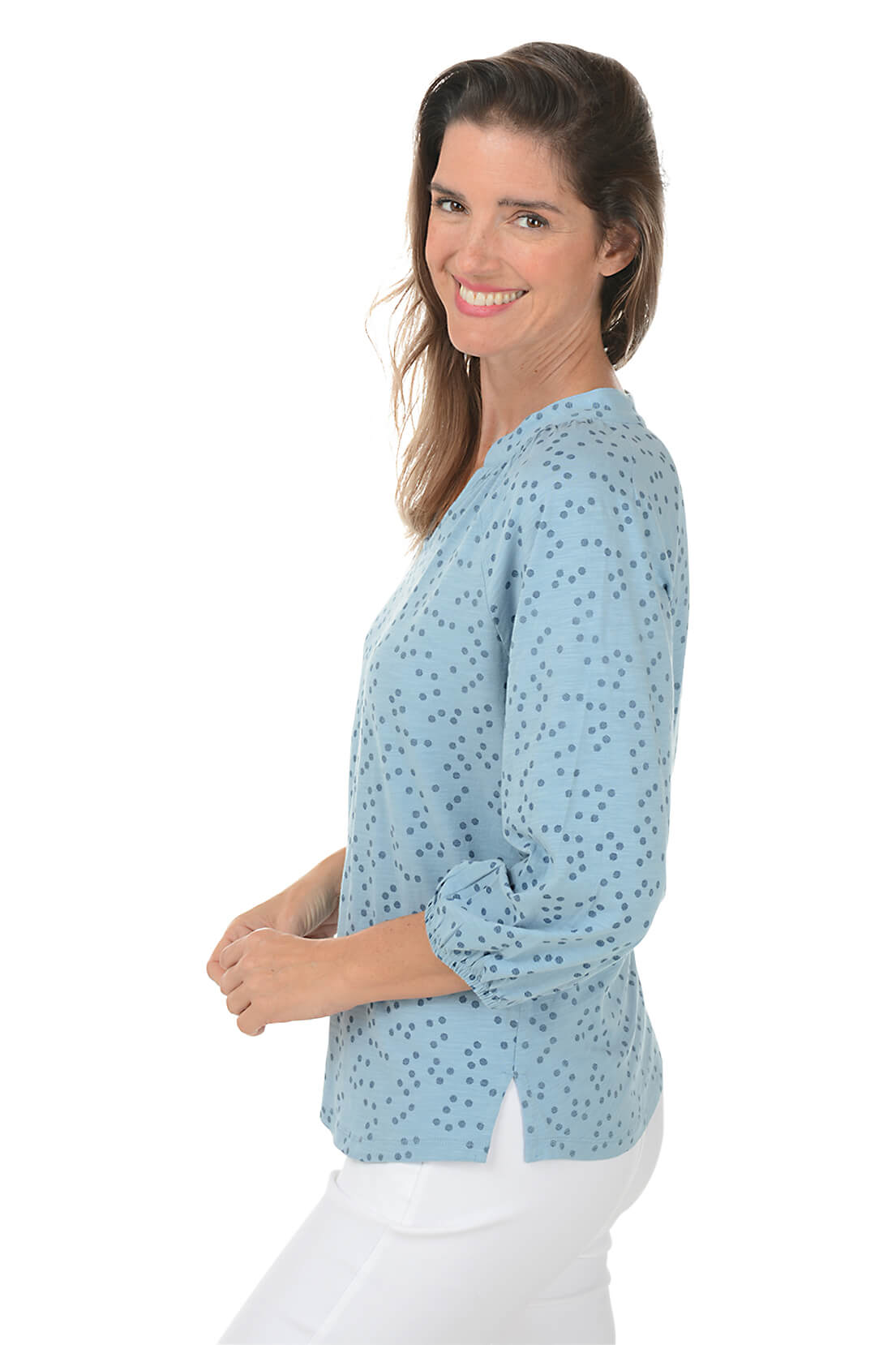A side shot of a woman wearing a blue dotted split neck slub knit top with 3/4 raglan sleeves and white pants.