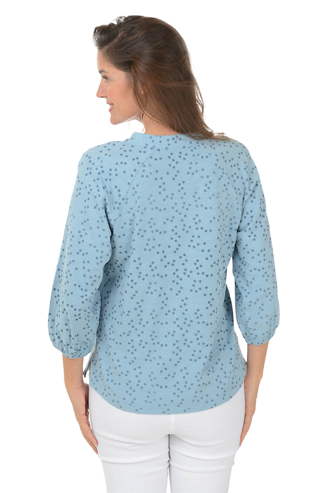 A back shot of a woman wearing a blue dotted split neck slub knit top with 3/4 raglan sleeves and white pants.