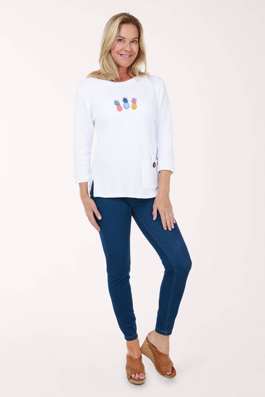 A woman wearing medium wash skinny pull-on jeggings with a white pineapple-print sweater and tan wedge sandals, shown full length.