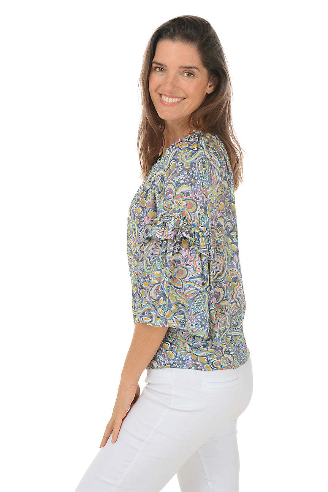 A side shot of a model wearing a floral paisley ruffled bell sleeve blouse with a scoop neck, in a multi-colored pattern. She is also wearing white pants.