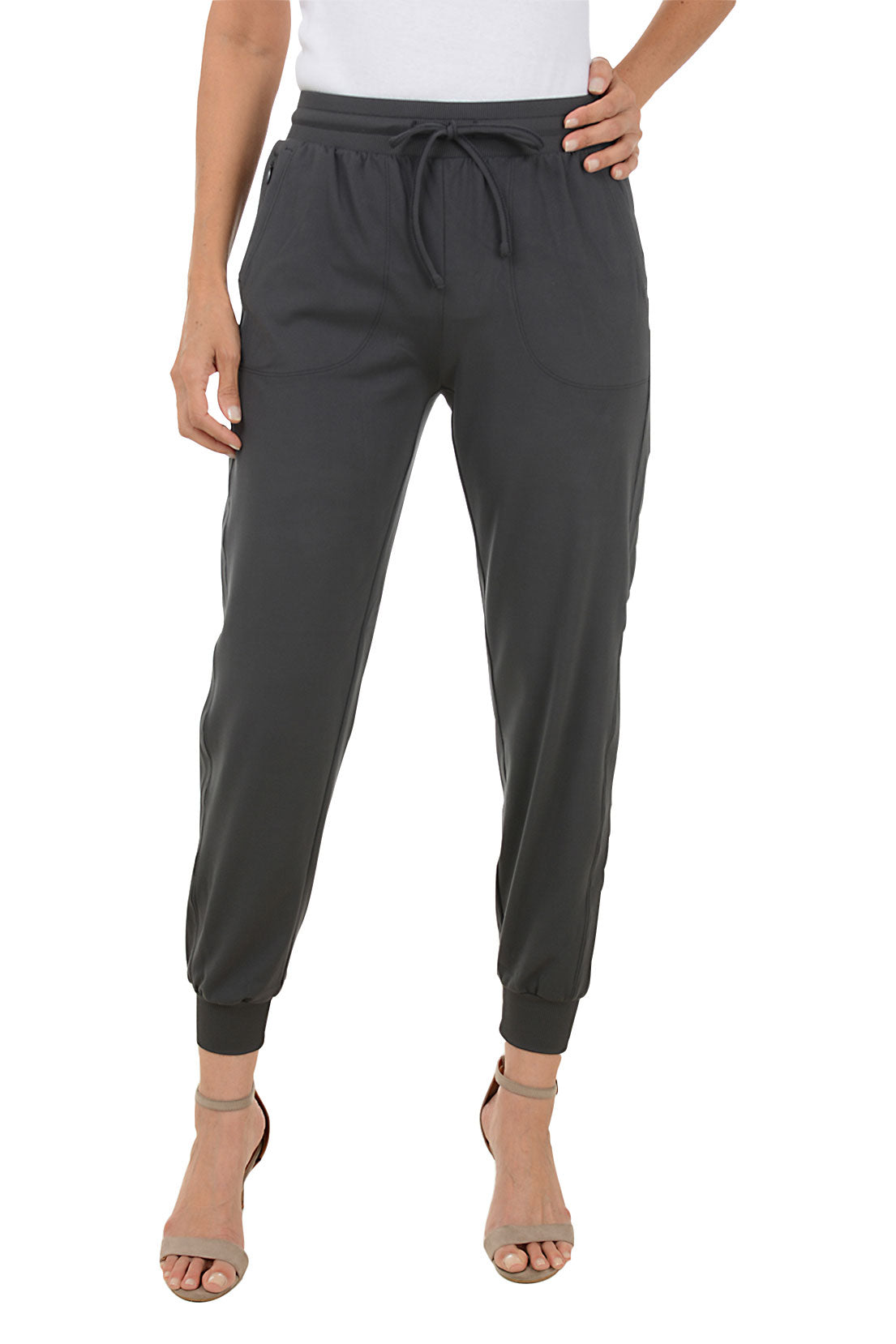 A front shot of a model wearing a pair of charcoal grey ankle-length jogger pants with an elastic waistband and adjustable drawstring, featuring front patch pockets with a zippered coin pocket, flat back, relaxed fit through the leg, and rib-knit cuffs at the ankle.