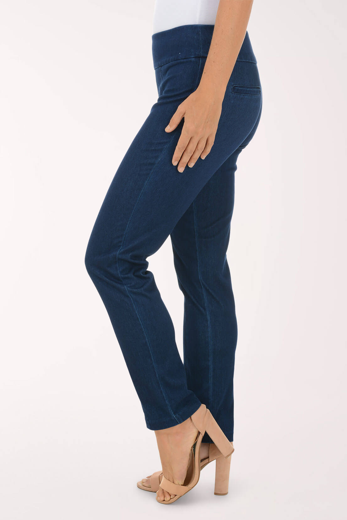A side view of dark wash slim leg pull-on jeans showing the slim full-length leg, worn with nude heels.