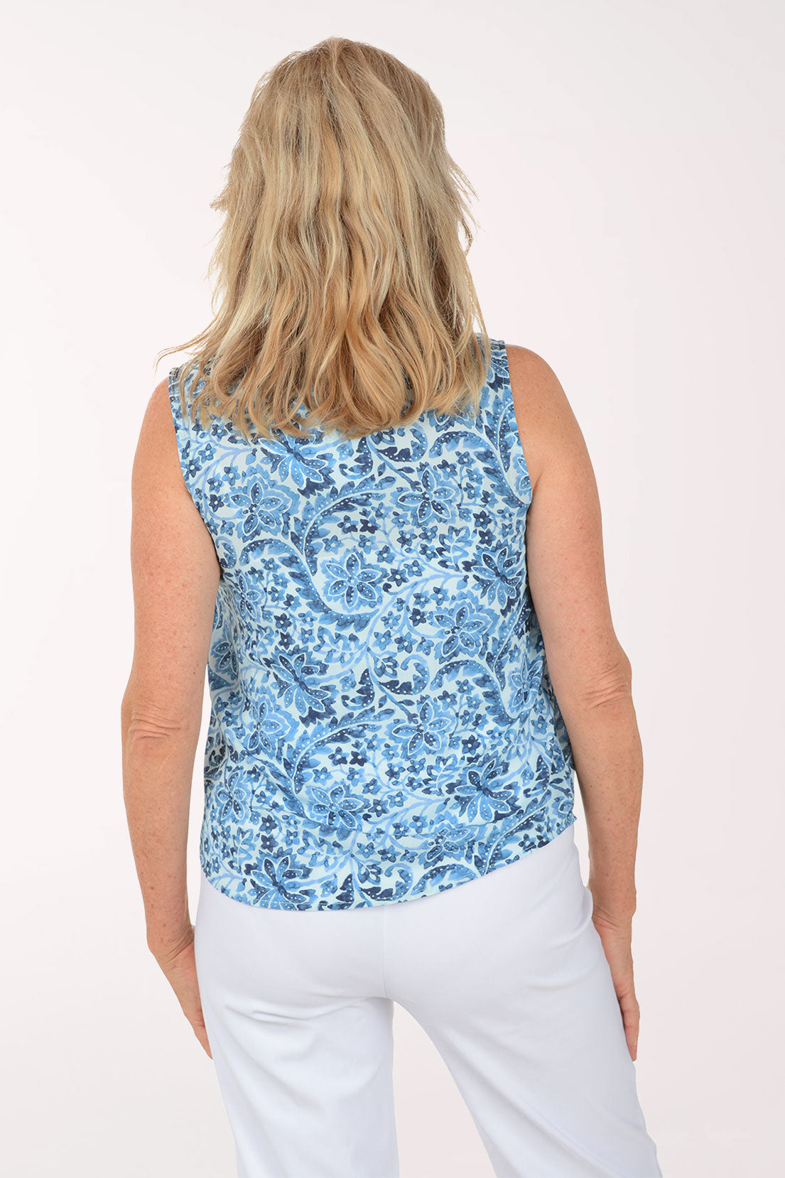 Woman wearing a blue floral sleeveless top and white pants on a white background