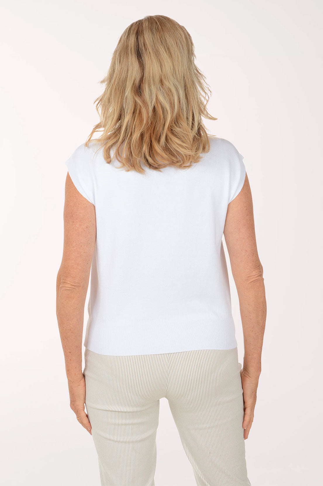 Woman wearing a white sleeveless sweater and light-colored pants on a plain background