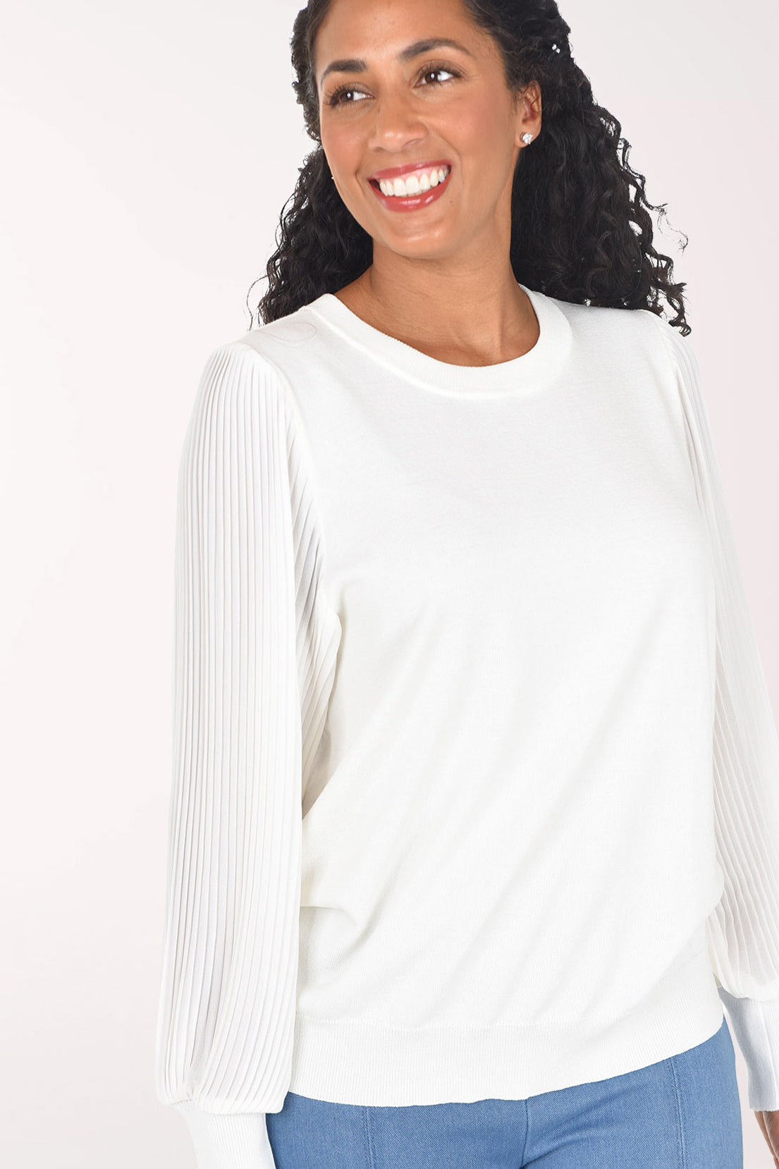 A woman modeling a white long-sleeve crewneck sweater with textured, pleated sheer sleeves, paired with blue jeans, against a plain light background.