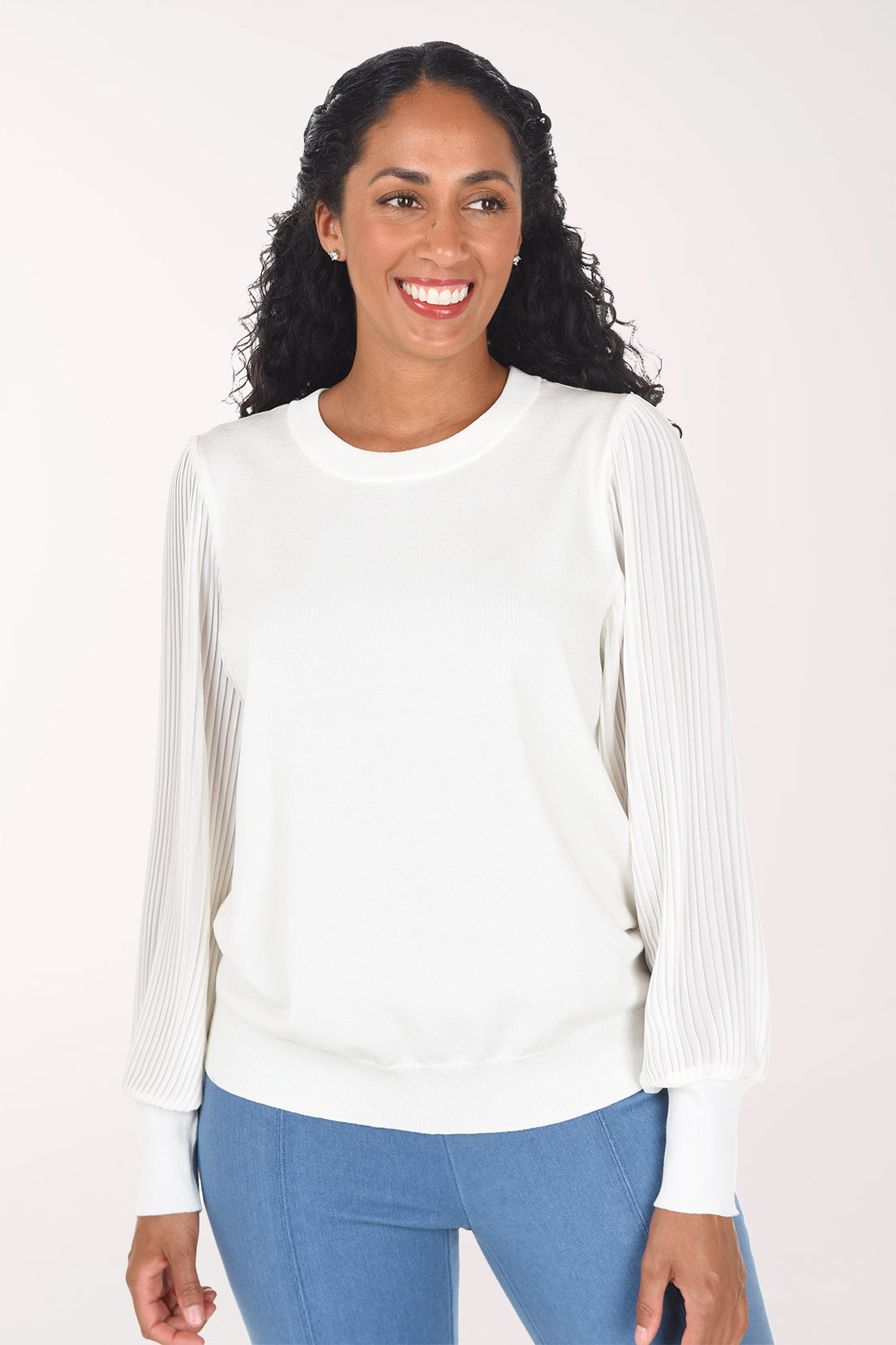 A woman modeling a white long-sleeve crewneck sweater with textured, pleated sheer sleeves, paired with blue jeans, against a plain light background.